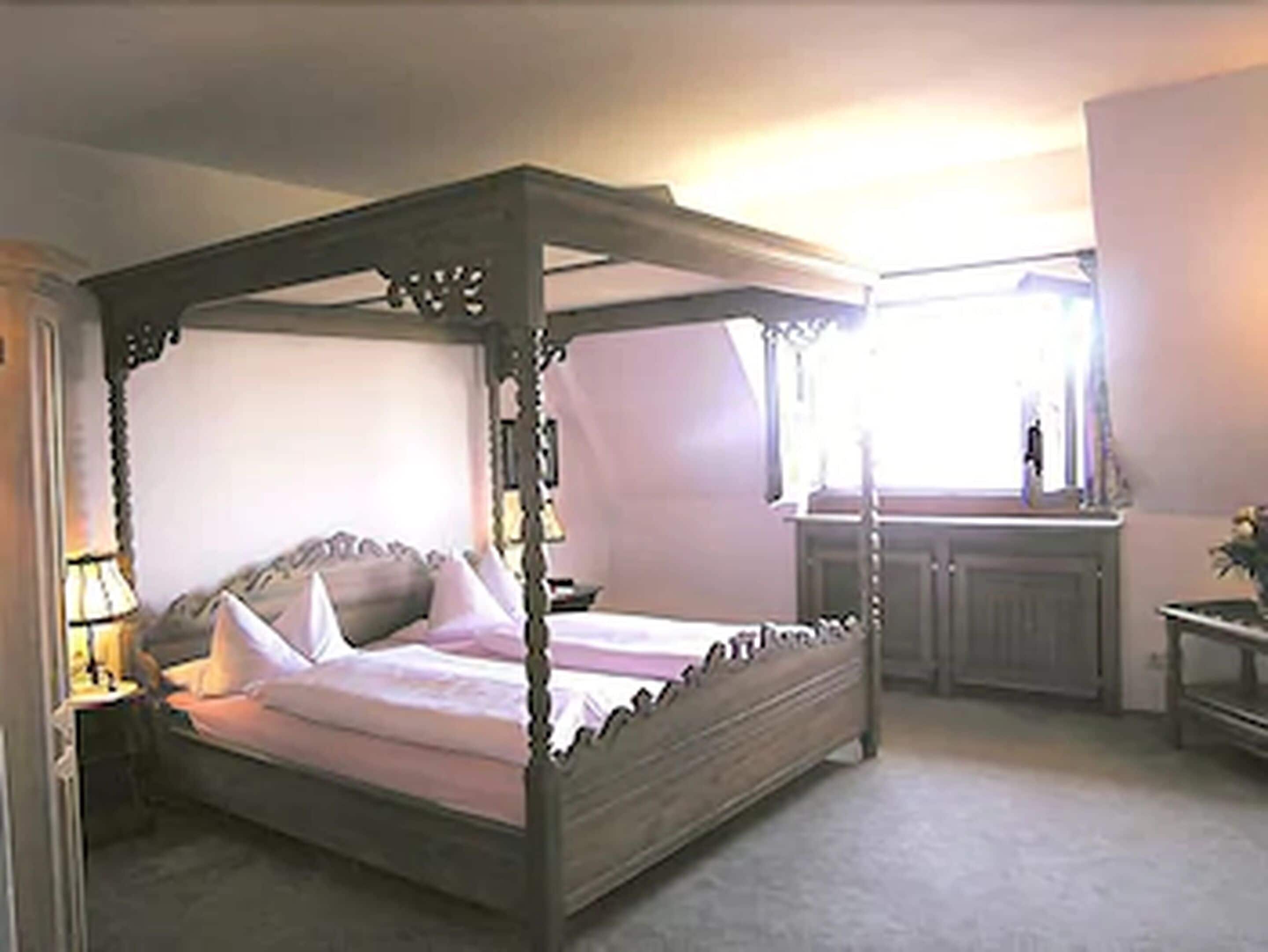 Una cama con dosel sobre cuatro columnas, un marco de madera, una mesita de noche con lámpara y una ventana con cortinas.