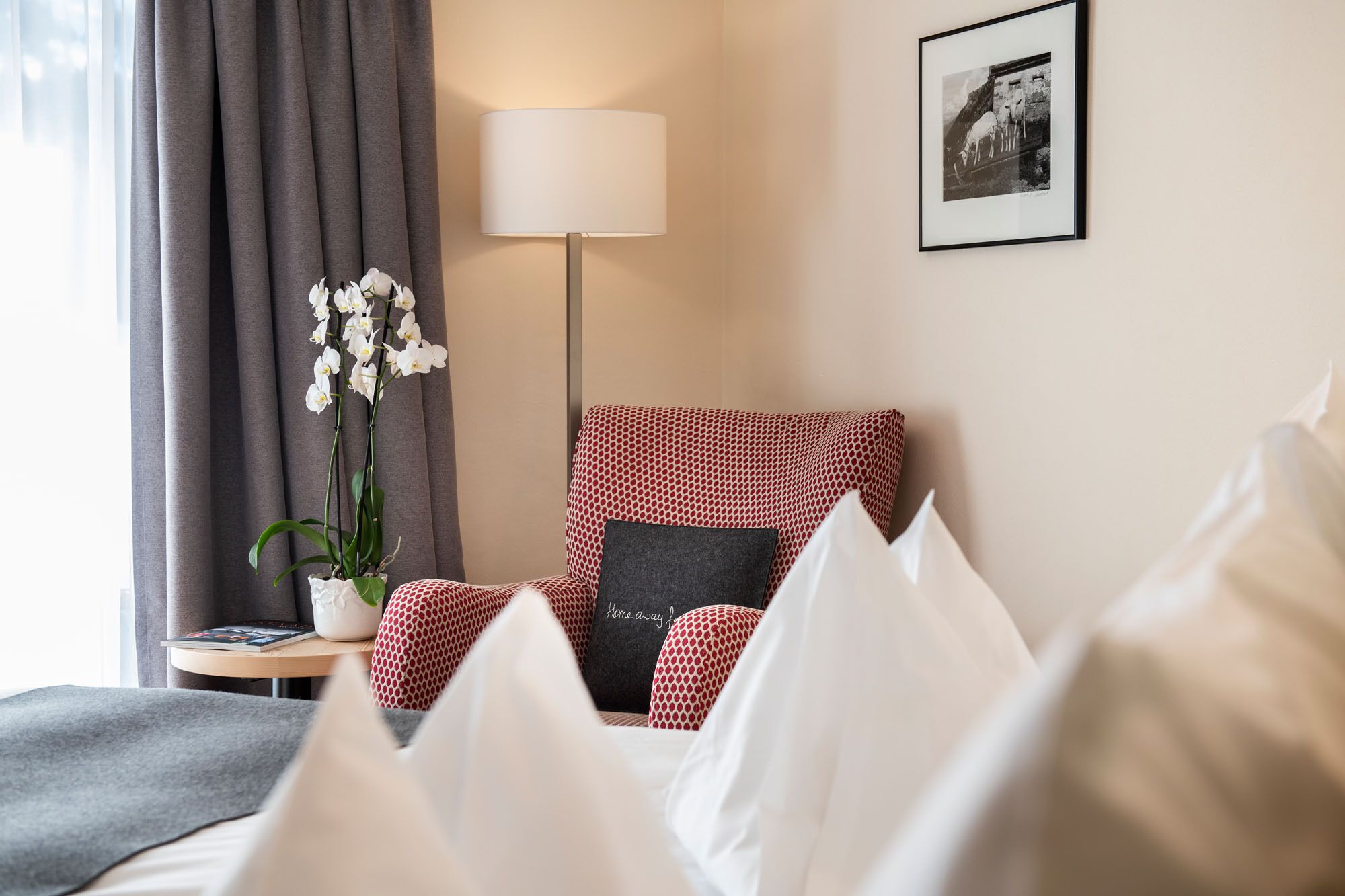 Quarto de hotel com cama, poltrona estampada em vermelho e branco, luminária de chão, quadro na parede e uma orquídea em vaso.