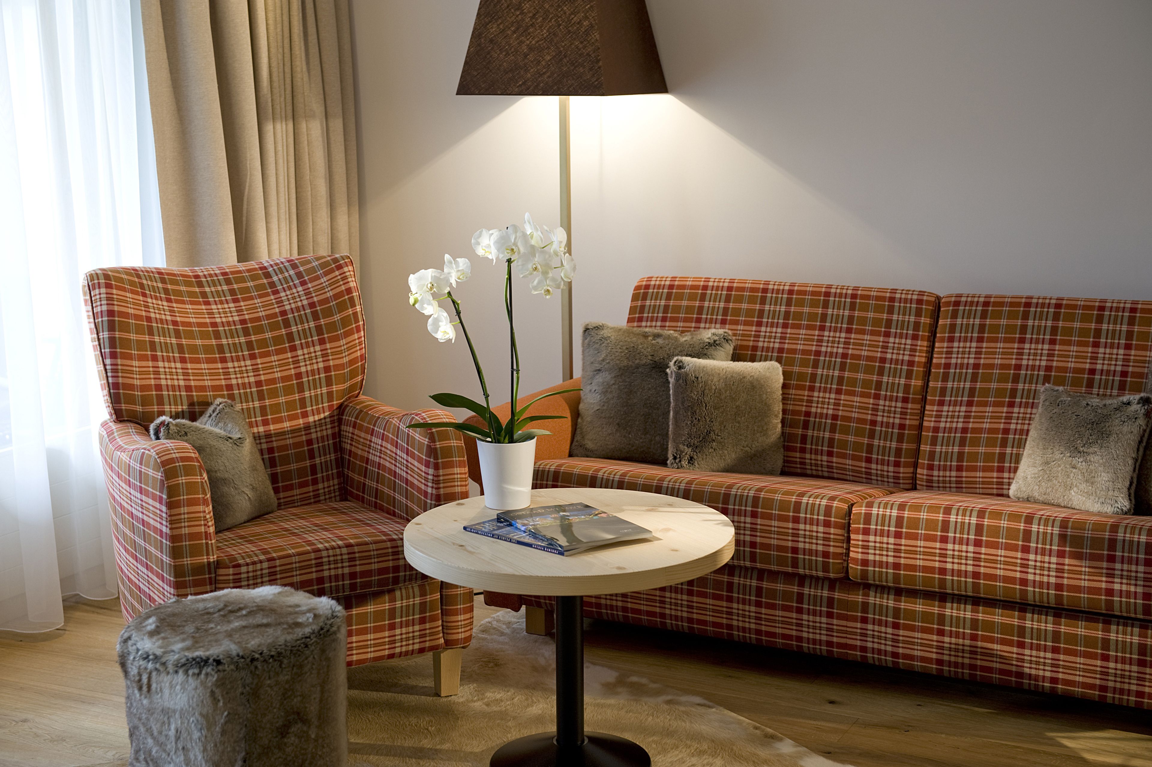 A living room with a plaid sofa, a matching armchair, a small round table with a book, a potted plant, and a lamp.