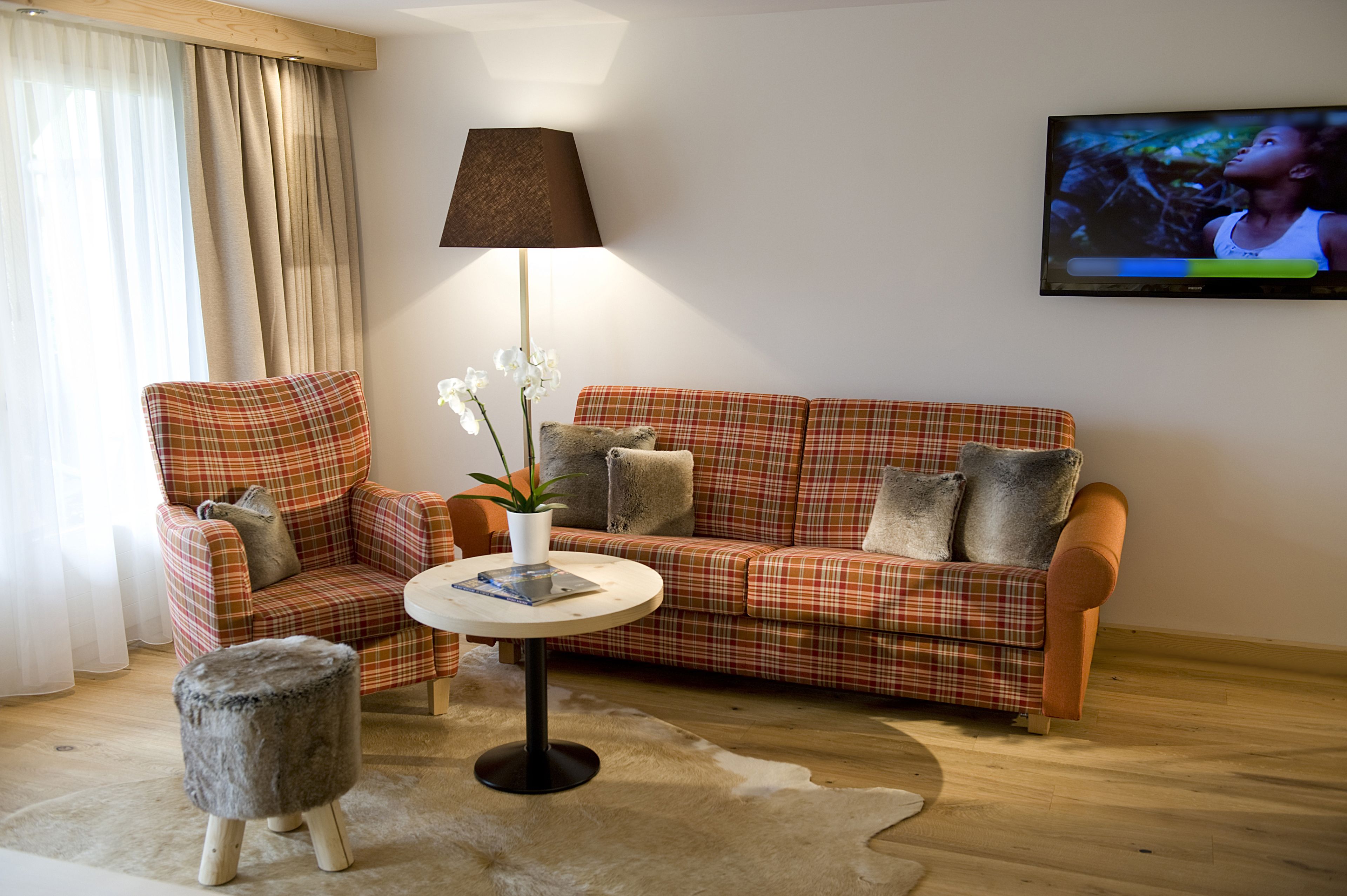 A living room with a plaid sofa, a matching armchair, a small round table with a lamp, and a television mounted on the wall.