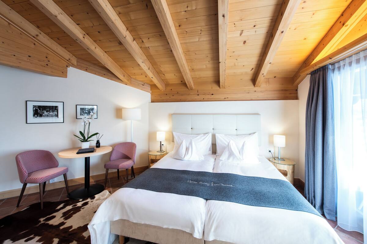 A hotel room with a wooden ceiling, a large bed with white bedding, two side tables with lamps, a small round table with a chair, and a cowhide rug.