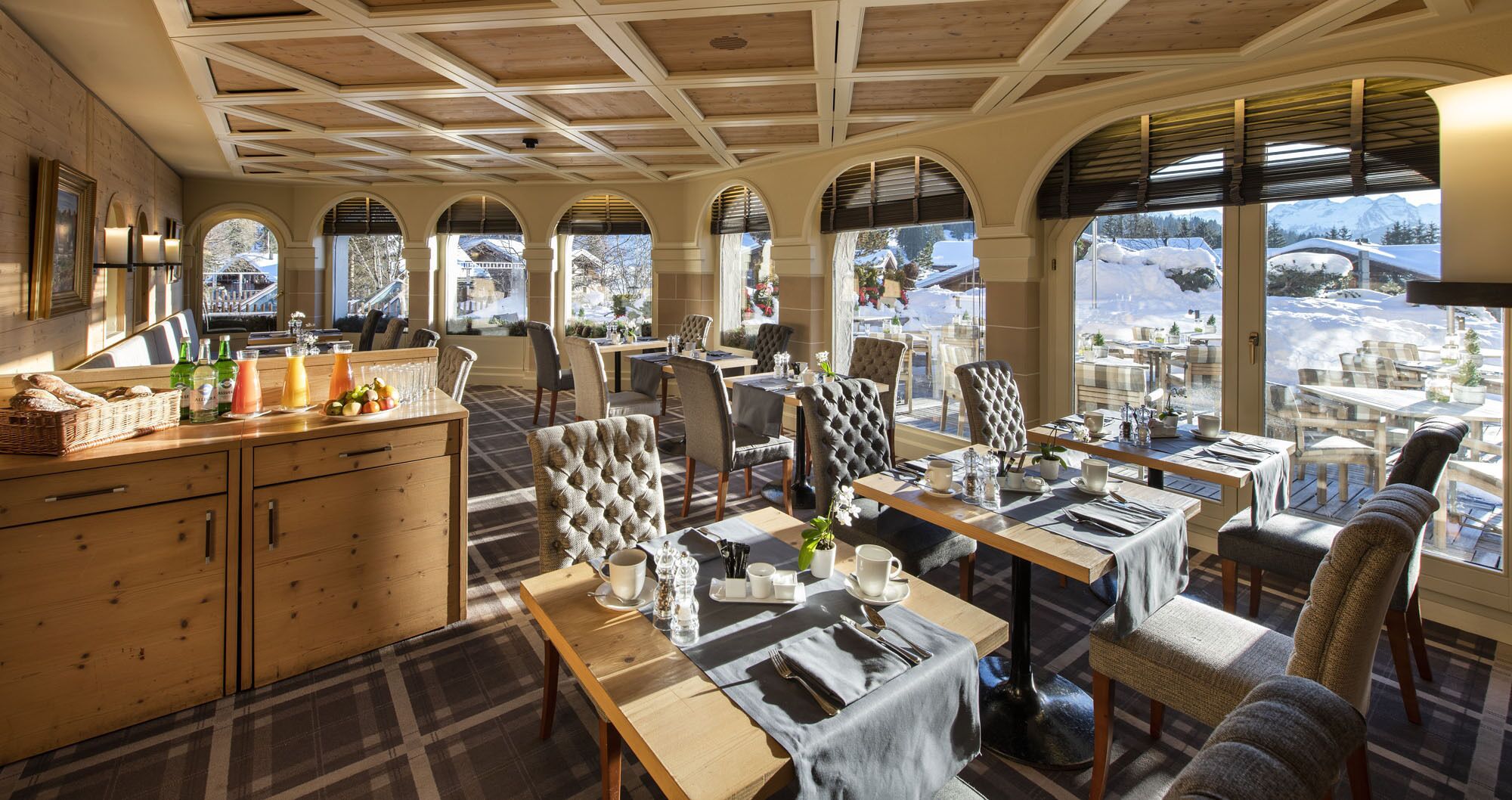 Restaurante con techo alto, vigas de madera y grandes ventanales con vistas a la montaña. Las mesas están dispuestas con manteles blancos y cubertería.