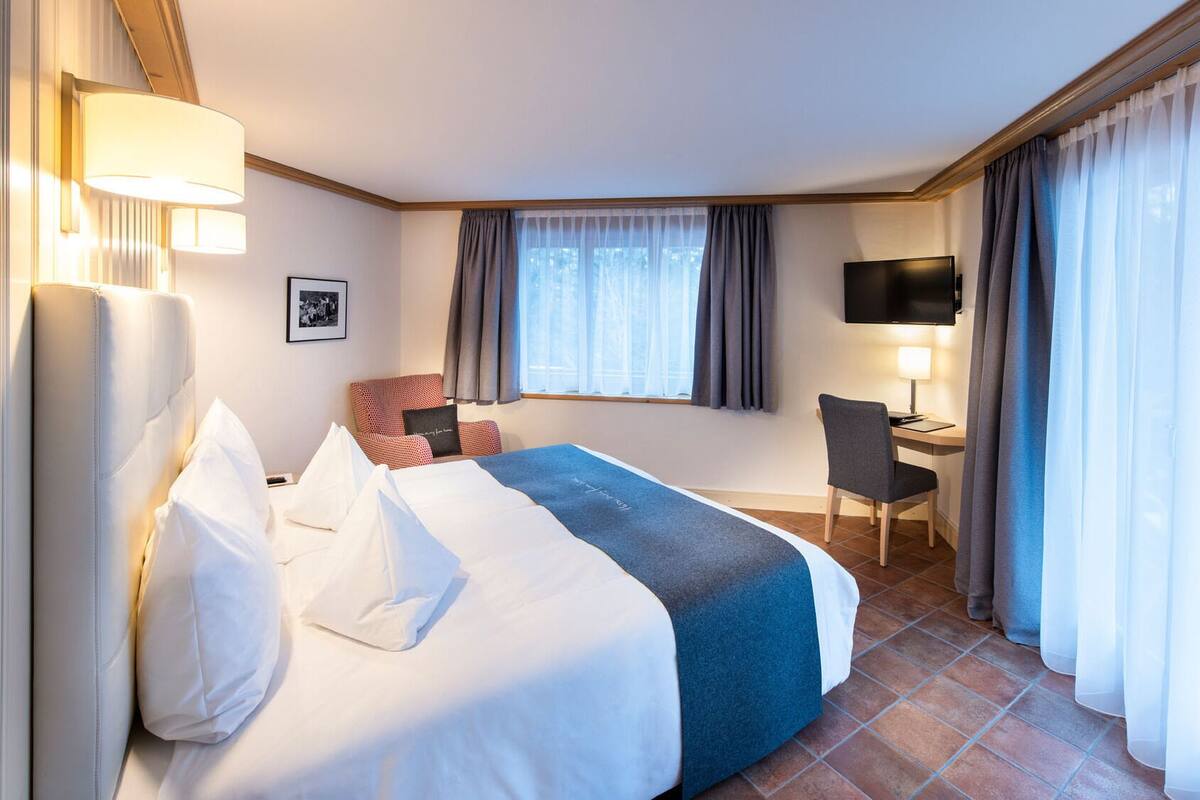 A hotel room with a bed, a desk, a chair, a TV, and a window with curtains.