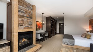 A modern hotel room with a stone fireplace, a bed with a gray and patterned blanket, a dining area with a table and chairs, and a hallway with a door.