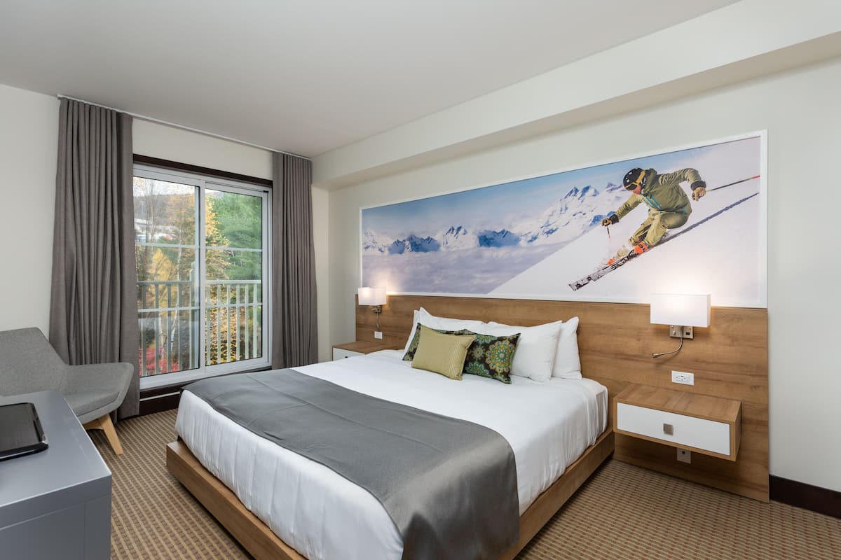 A hotel room with a large bed, a desk, a window with curtains, and a wall-mounted painting of a skier.