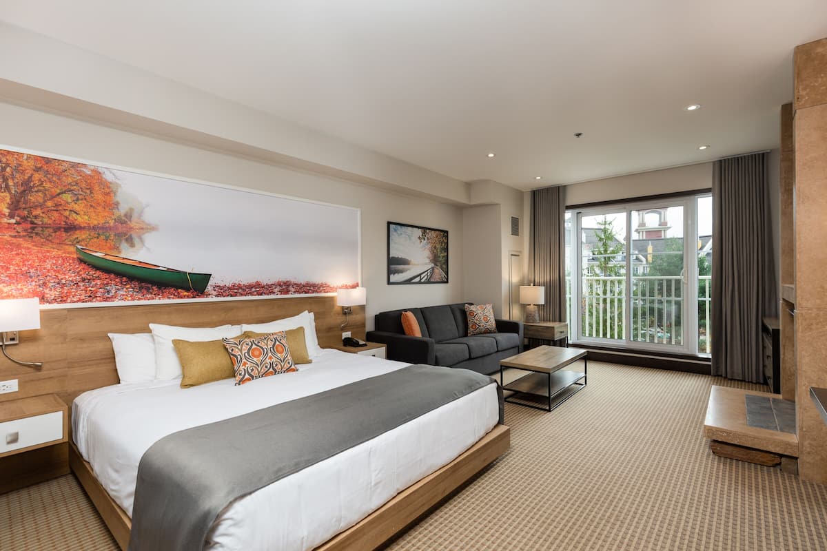 A modern hotel room with a large bed, a sofa, a coffee table, and a view of the outdoors through a large window.