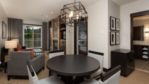 A modern hotel room with a dining area, a sofa, a lamp, a kitchen area, and a large window.