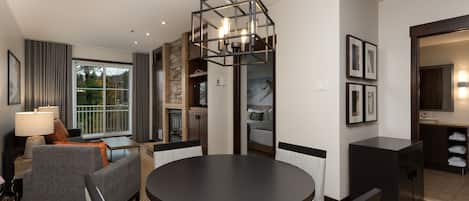 A modern hotel room with a dining area, a sofa, a lamp, a kitchen area, and a large window.