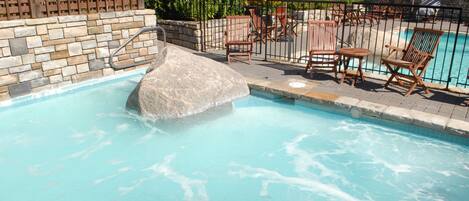 A swimming pool with a stone wall, wooden chairs, and a patio area.