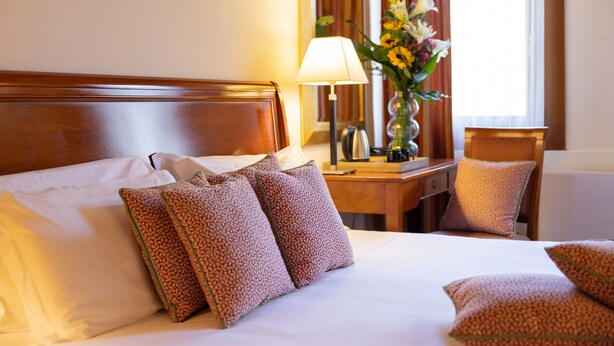 Ein Hotelzimmer mit einem hölzernen Kopfteil, einem Bett mit weißen Bettwäsche und gemusterten Kissen, einem Nachttisch mit Lampe und Blumenvase sowie einem Sessel mit Sitzkissen.