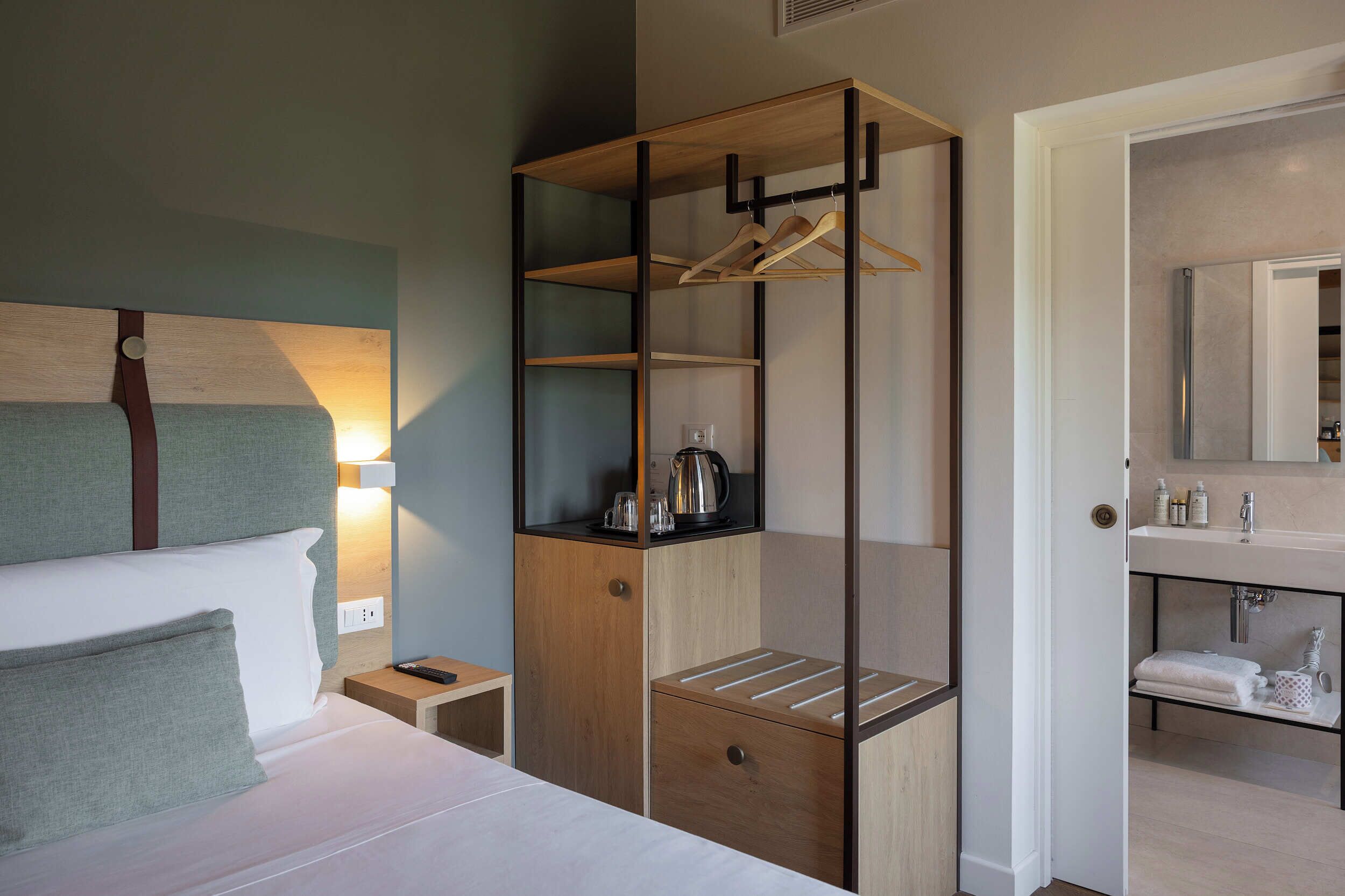 A hotel room with a bed, a wooden wardrobe with shelves, a nightstand, and a bathroom with a sink and mirror.