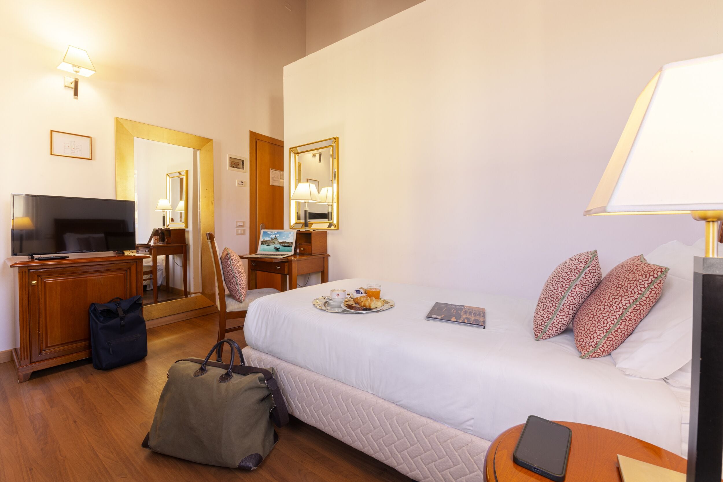 A hotel room with a bed, bedside table, lamp, desk with a computer, chair, and a TV.
