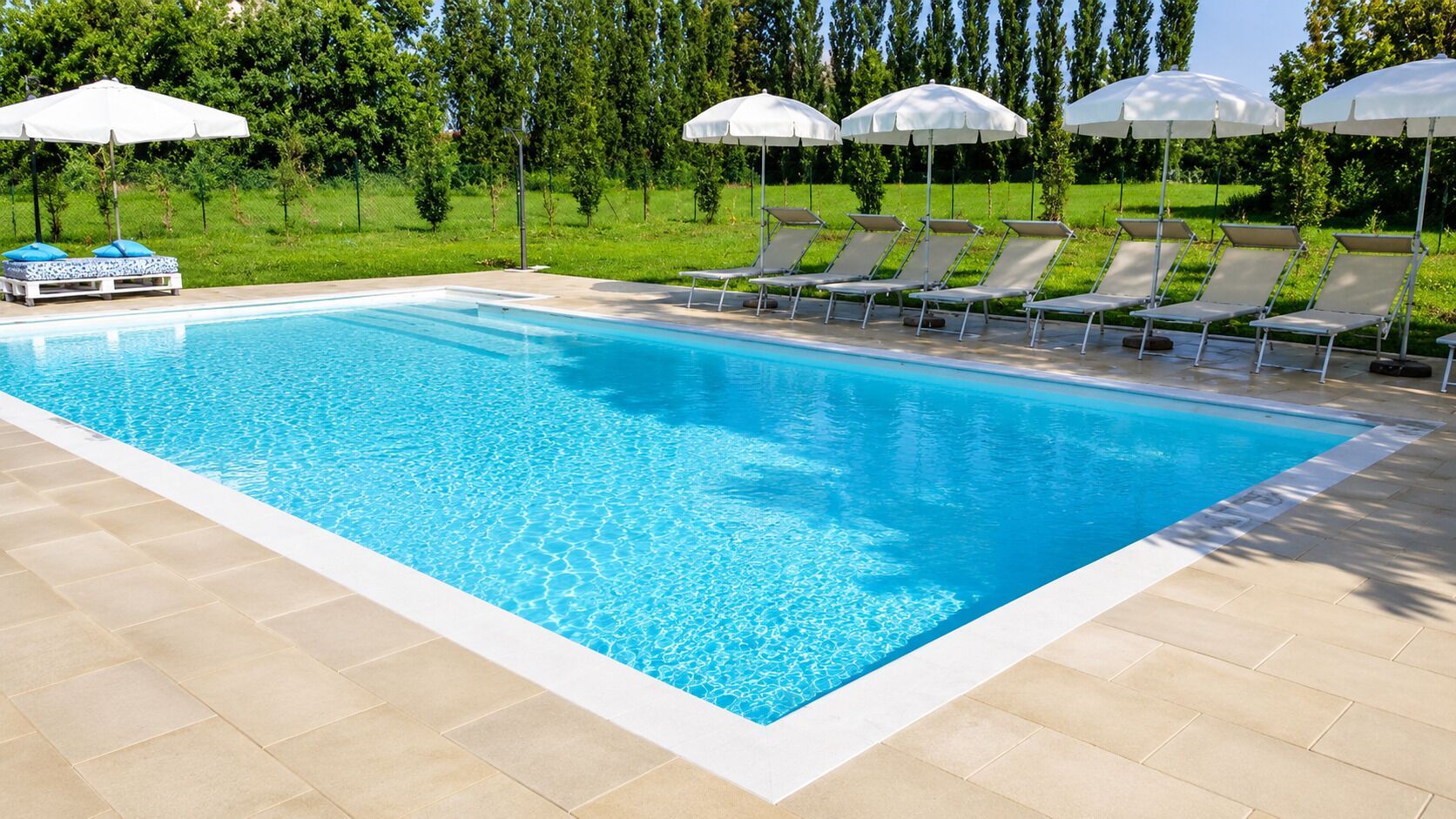 Una piscina rectangular con agua azul cristalina, bordeada por baldosas color beige. Hay sombrillas blancas y sillones de descanso con cojines azules.