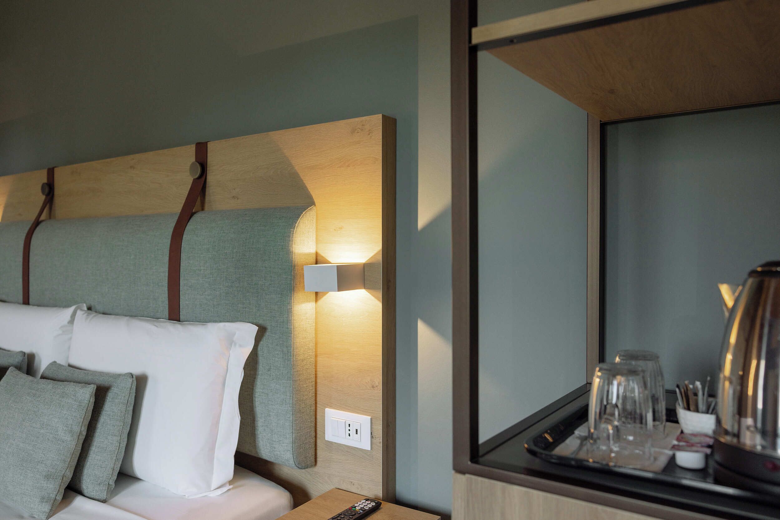 A hotel room with a bed, pillows, a bedside table with a remote control, and a tray with glasses and a kettle.