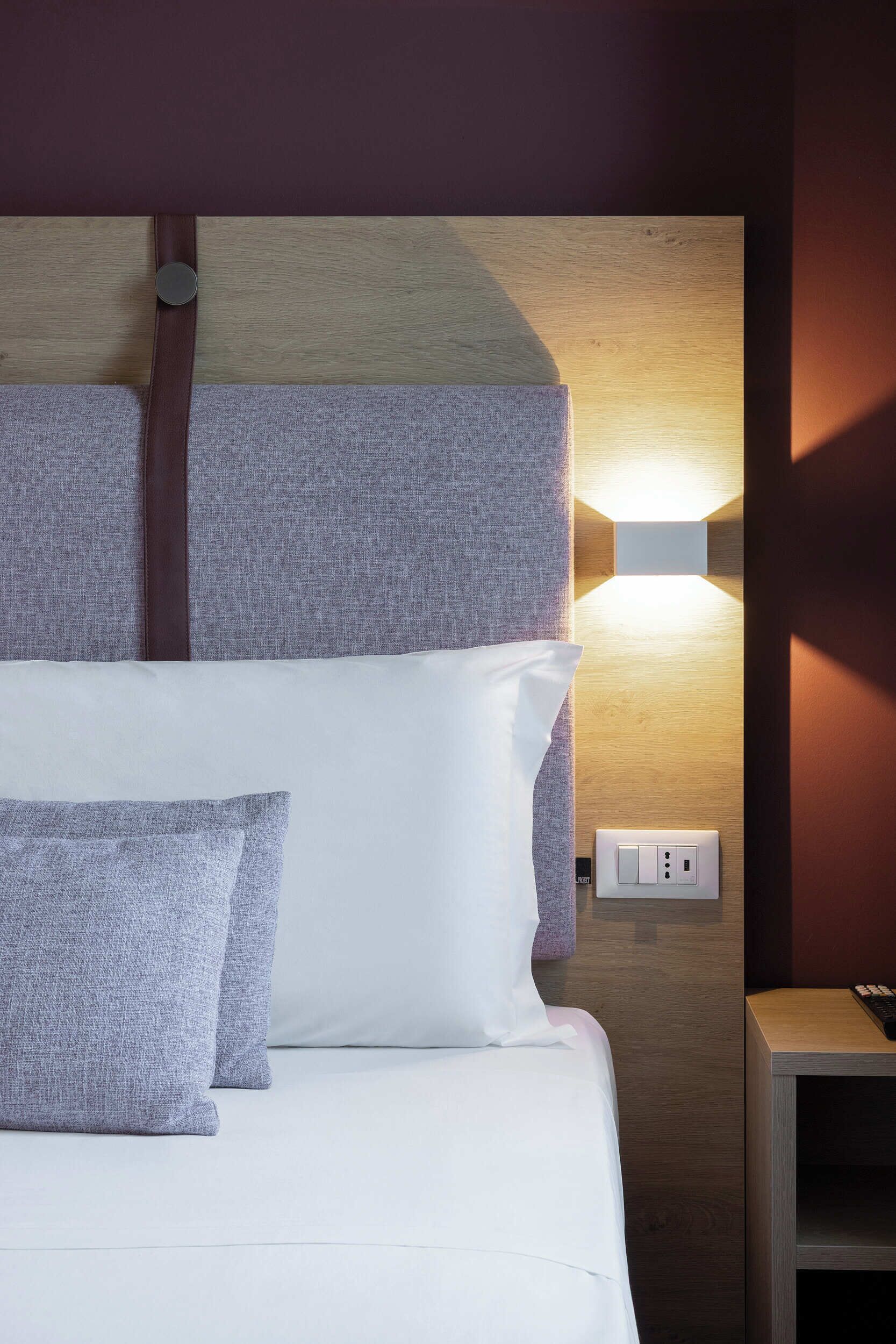 Una cama bien tendida con ropa de cama blanca y gris, una lámpara fijada a la pared y un enchufe.