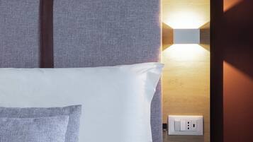 Una cama bien tendida con ropa de cama blanca y gris, una lámpara fijada a la pared y un enchufe.