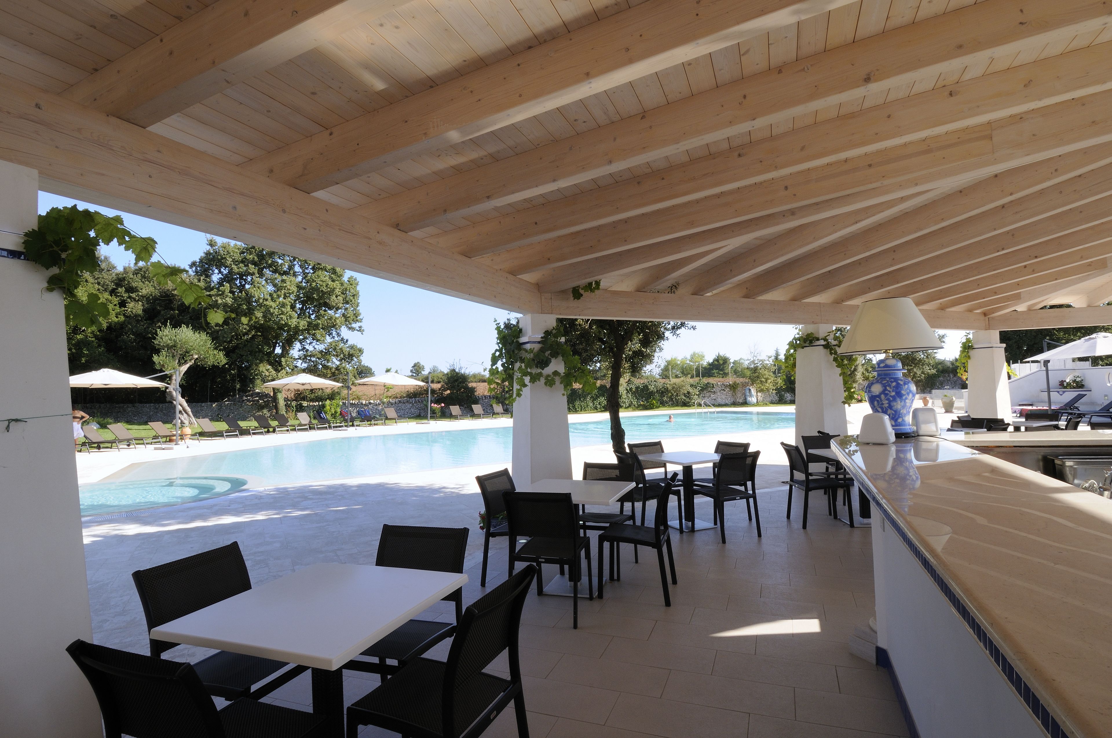 An outdoor pool area with lounge chairs and tables, a wooden ceiling, and a view of the pool.