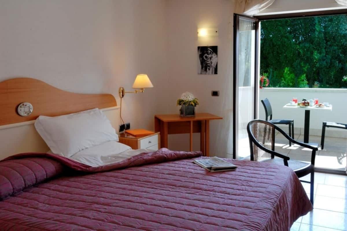 Una habitación de hotel con una cama, una mesita de noche, una lámpara, una pequeña mesa con un florero, una silla y vistas a zonas verdes por la ventana.