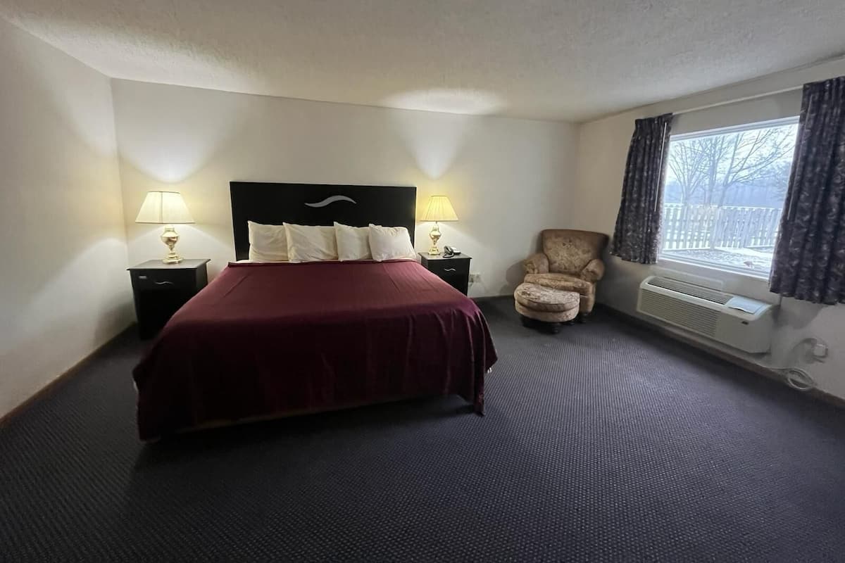 Habitación de hotel con una cama grande, dos mesitas de noche con lámparas, una silla, un ventanal con cortinas y un radiador.