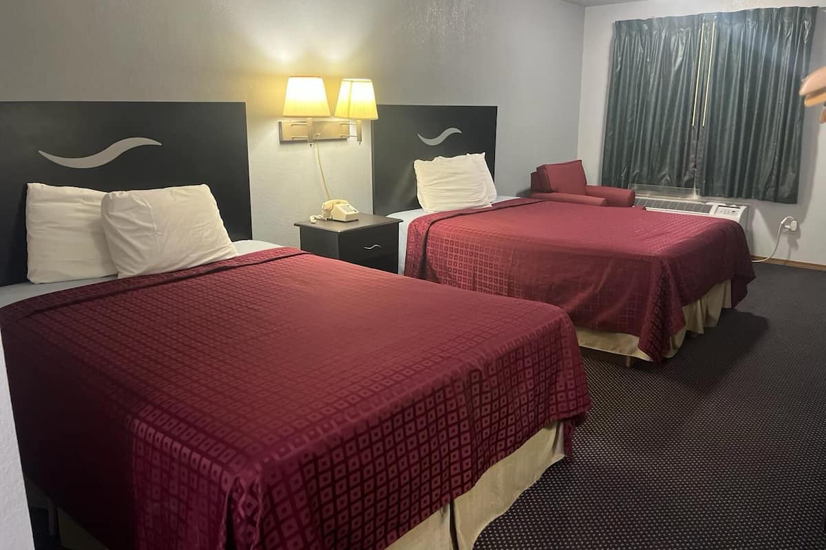 Habitación de hotel con dos camas, una mesita de noche con lámpara, teléfono y ventana con cortinas.