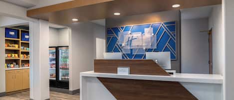 A reception desk with a modern design, a blue geometric wall decoration, and a refrigerator stocked with beverages.