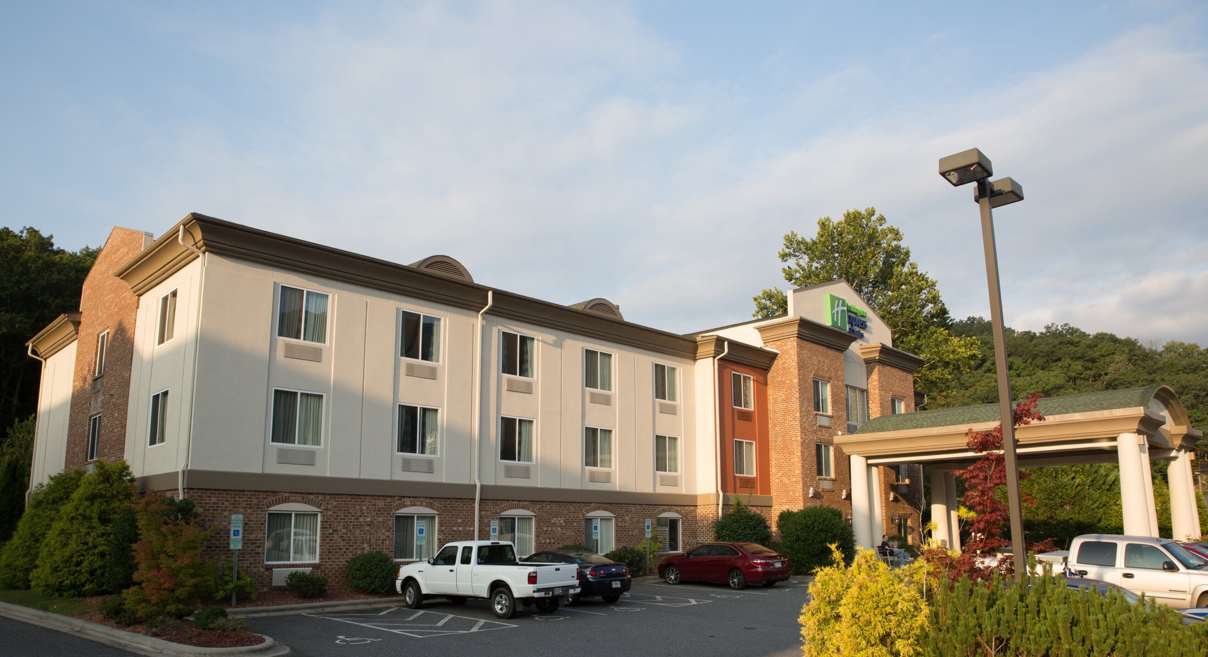 Un hotel de varios pisos con estacionamiento, un camino de acceso a la entrada y una zona ajardinada con arbustos.