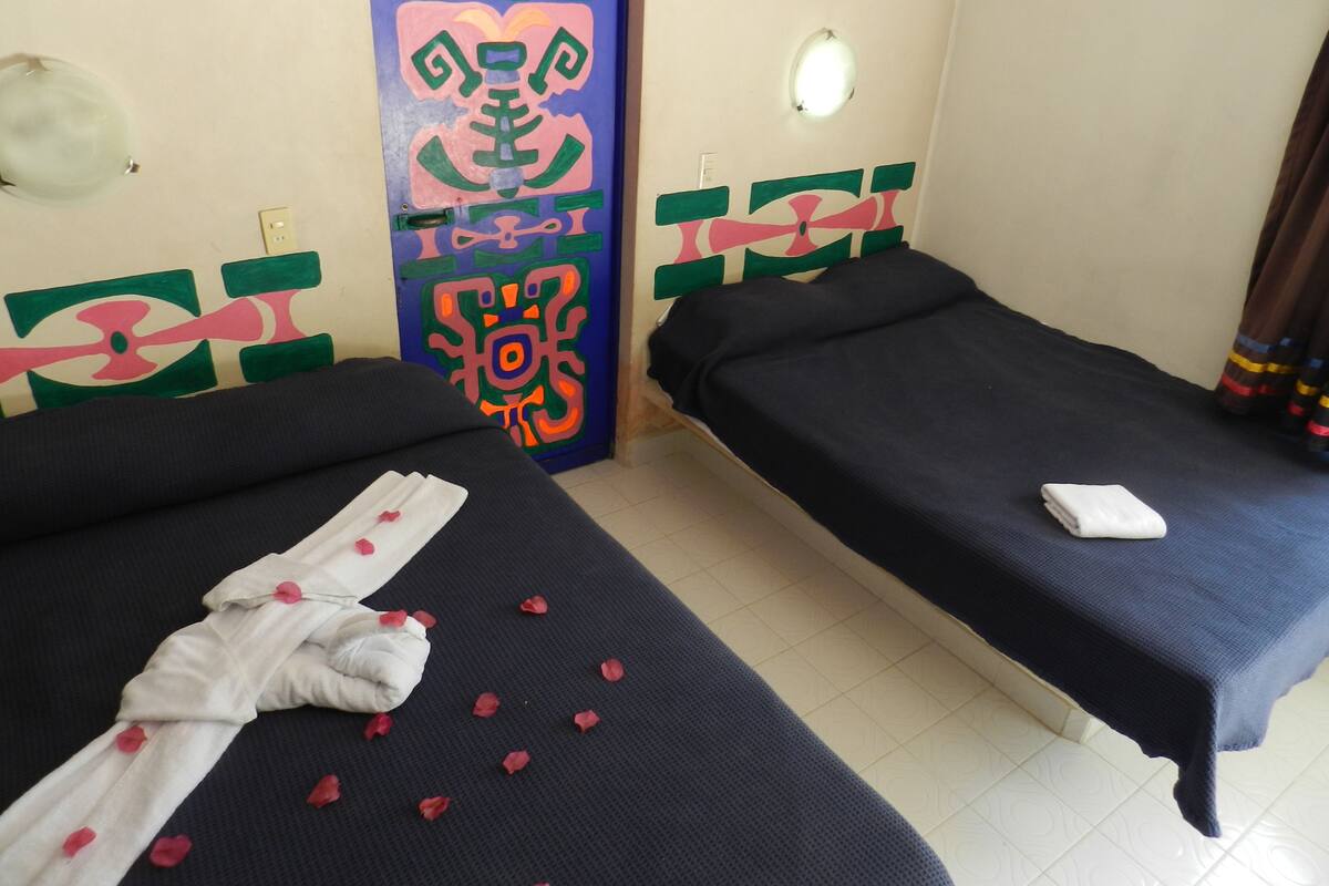 A hotel room with two beds, one with a towel and rose petals, and a colorful wall decoration.
