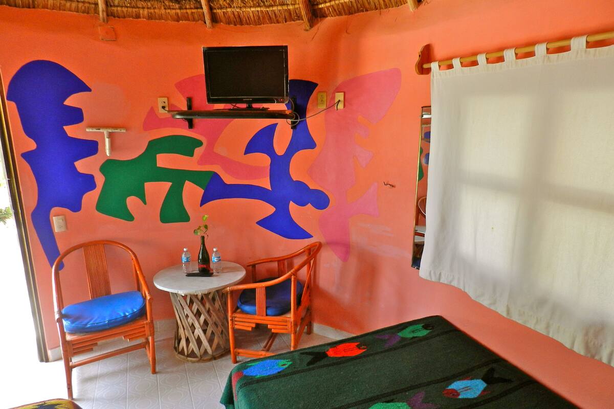 A room with two wicker chairs, a small round table, a bed with a green blanket, a television on a stand, and a wall with colorful abstract art.