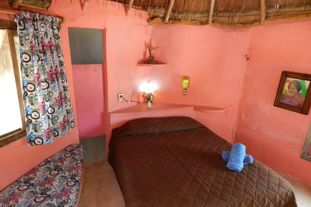 A small, pink-walled room with a single bed, a bench, a small shelf with a vase, and a framed picture on the wall.