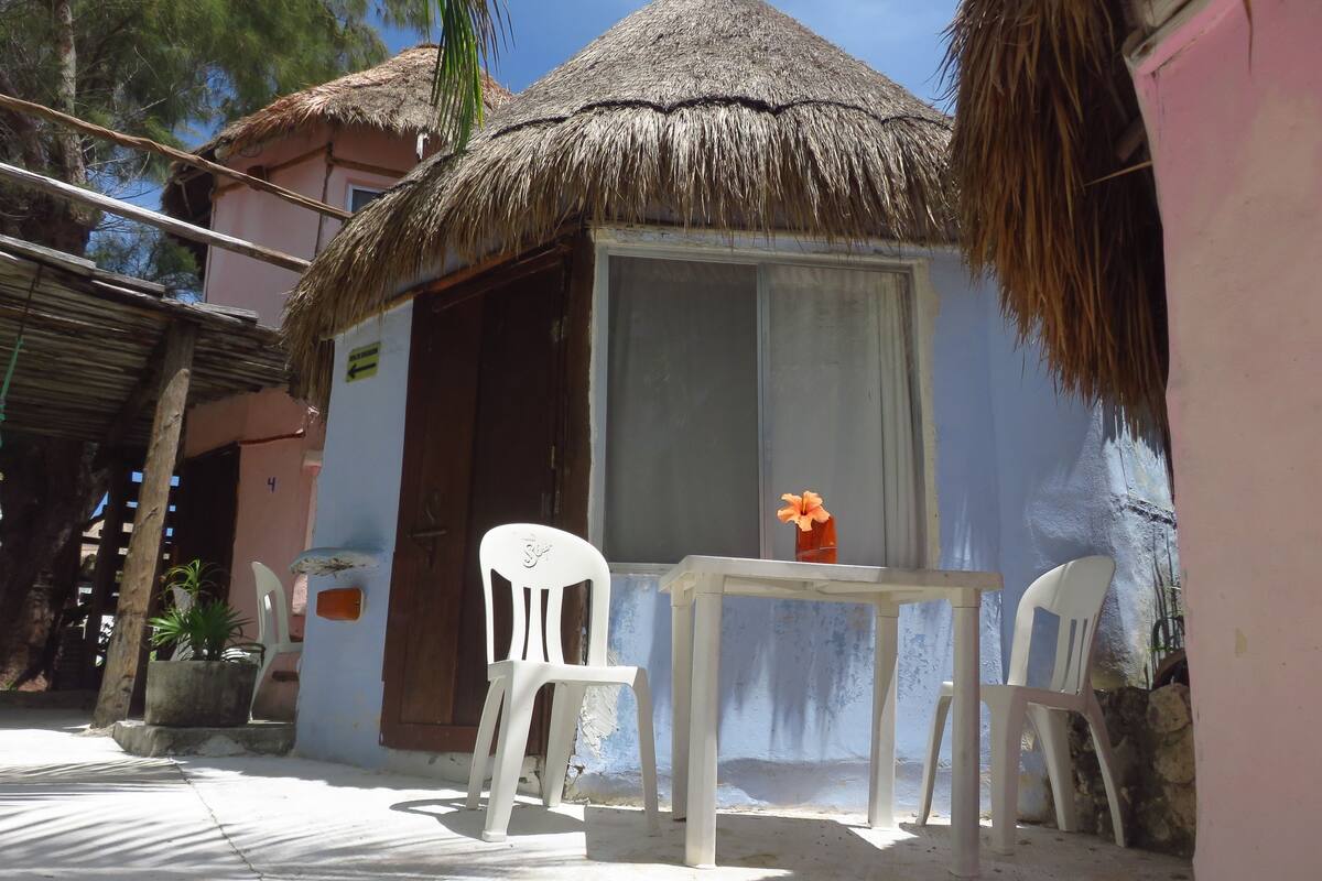 A small, single-room structure with a thatched roof, white plastic chairs, and a table with a vase of flowers.
