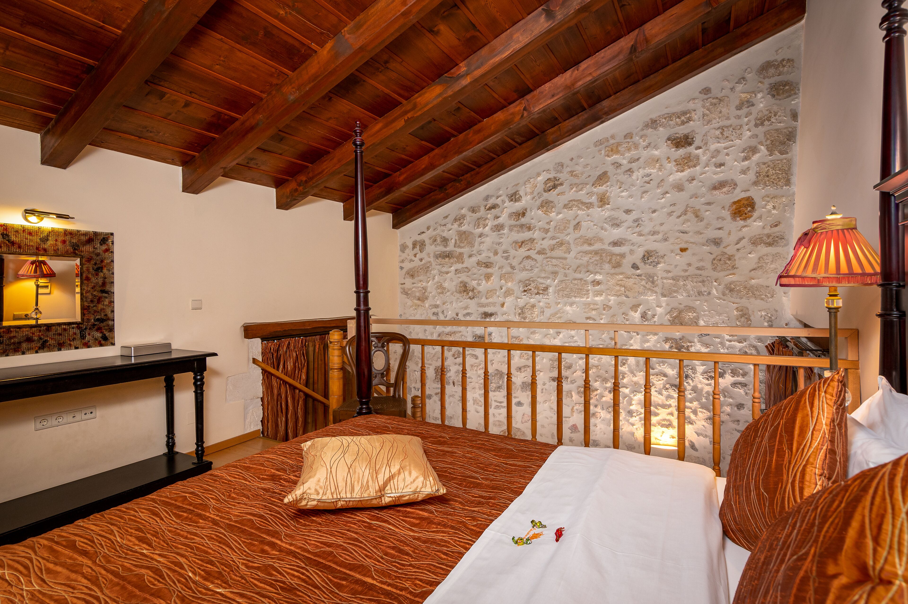 A bedroom with a stone wall, wooden ceiling beams, a bed with patterned bedding, a nightstand, a lamp, and a small table.