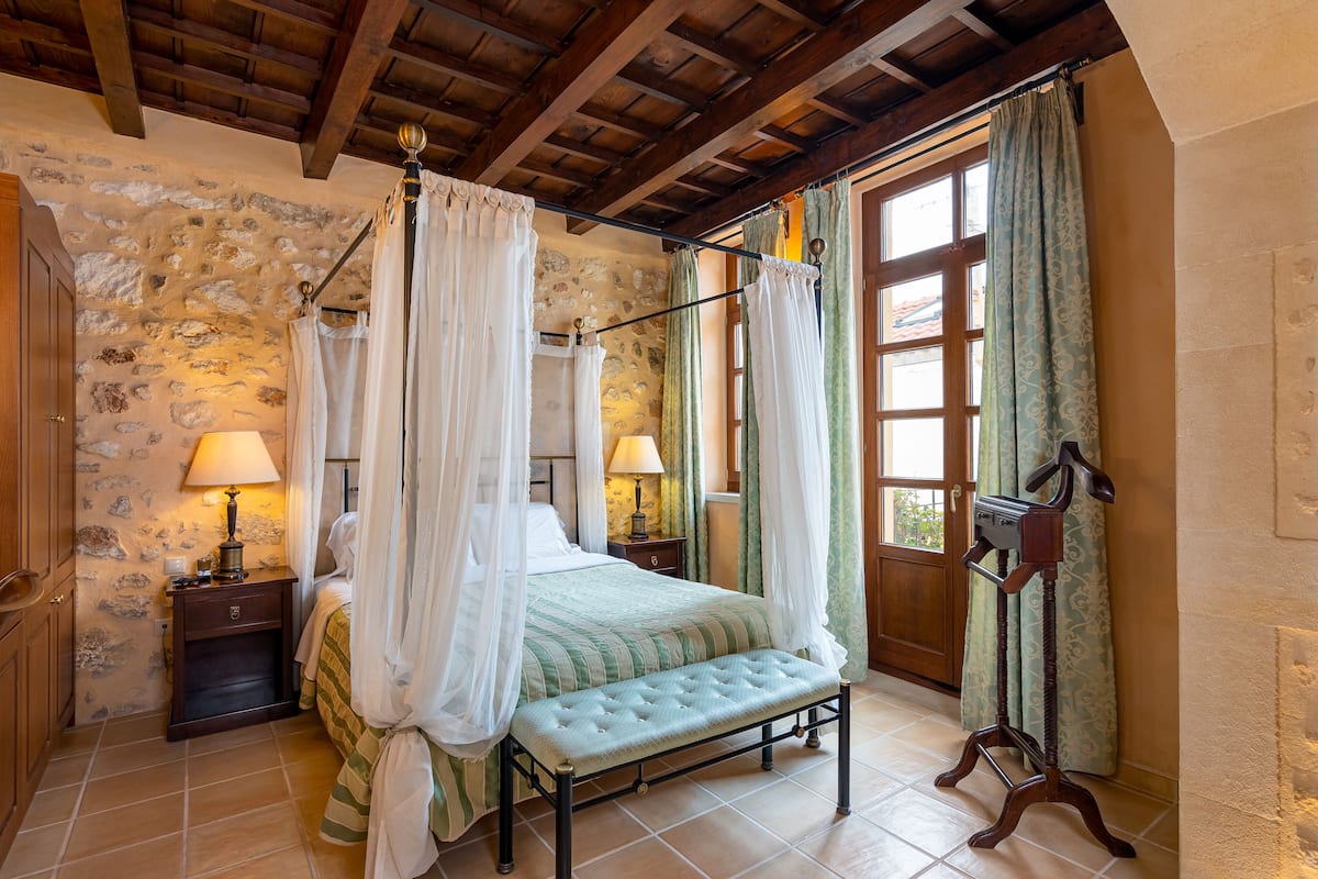 A bedroom with a stone wall, wooden ceiling beams, a four-poster bed with white curtains, a bedside table with a lamp, and a wooden bench.