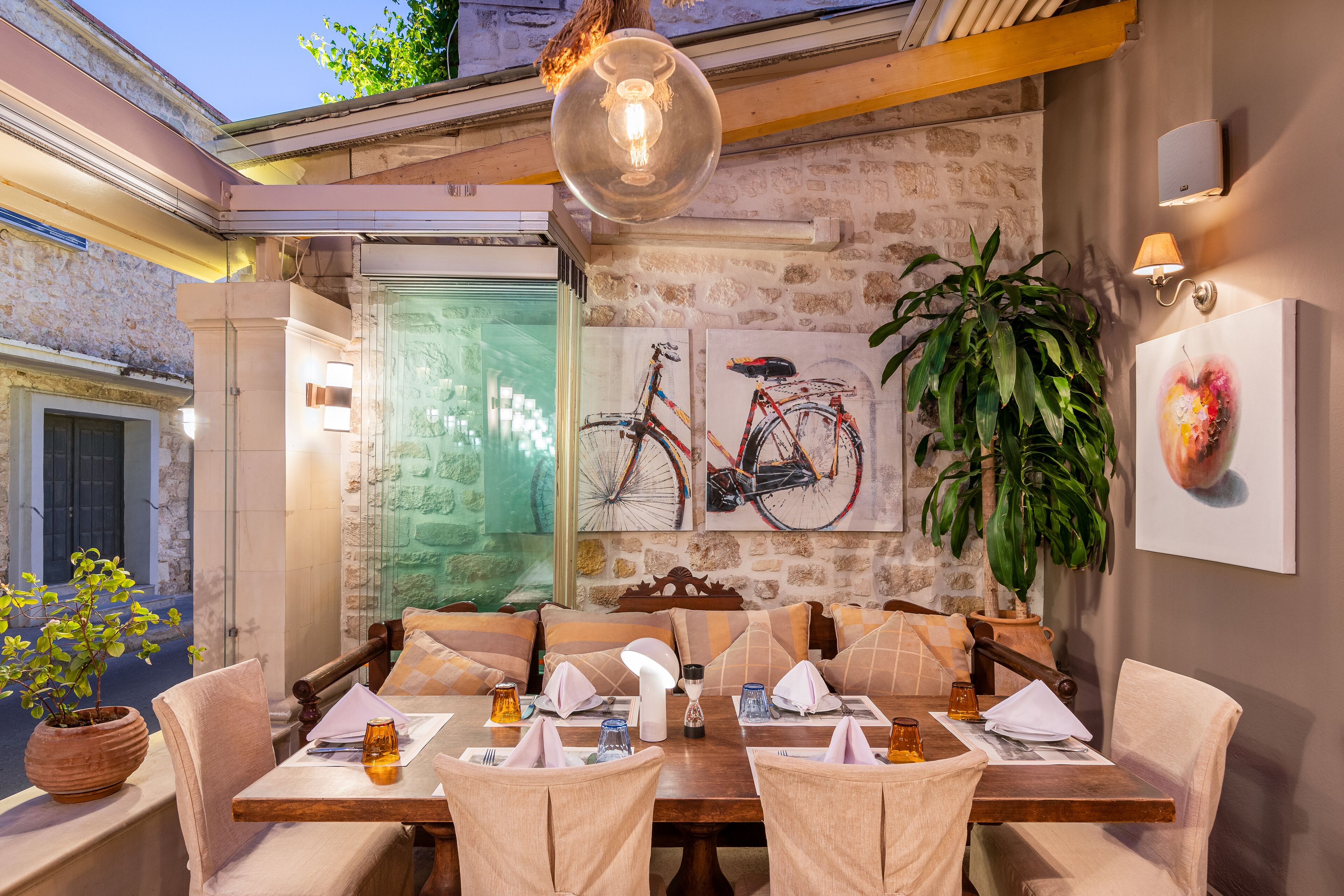 A dining area with a wooden table set for four, surrounded by cushioned chairs. There are two wall-mounted light fixtures and a potted plant to the left.