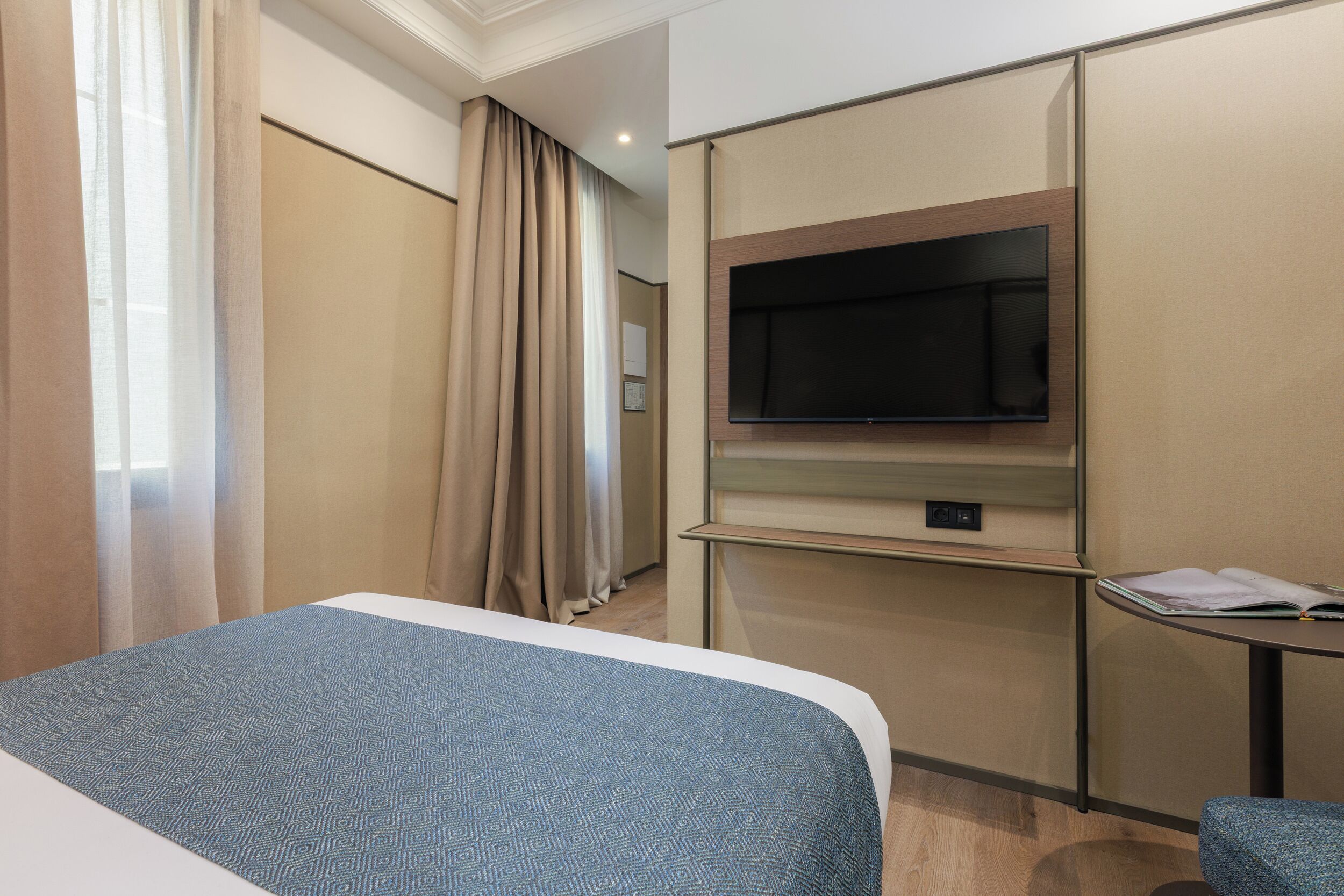 A hotel room with a bed, a TV mounted on the wall, a small table, and a window with curtains.