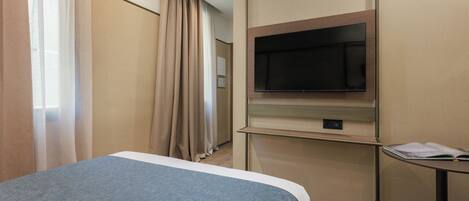 A hotel room with a bed, a TV mounted on the wall, a small table, and a window with curtains.