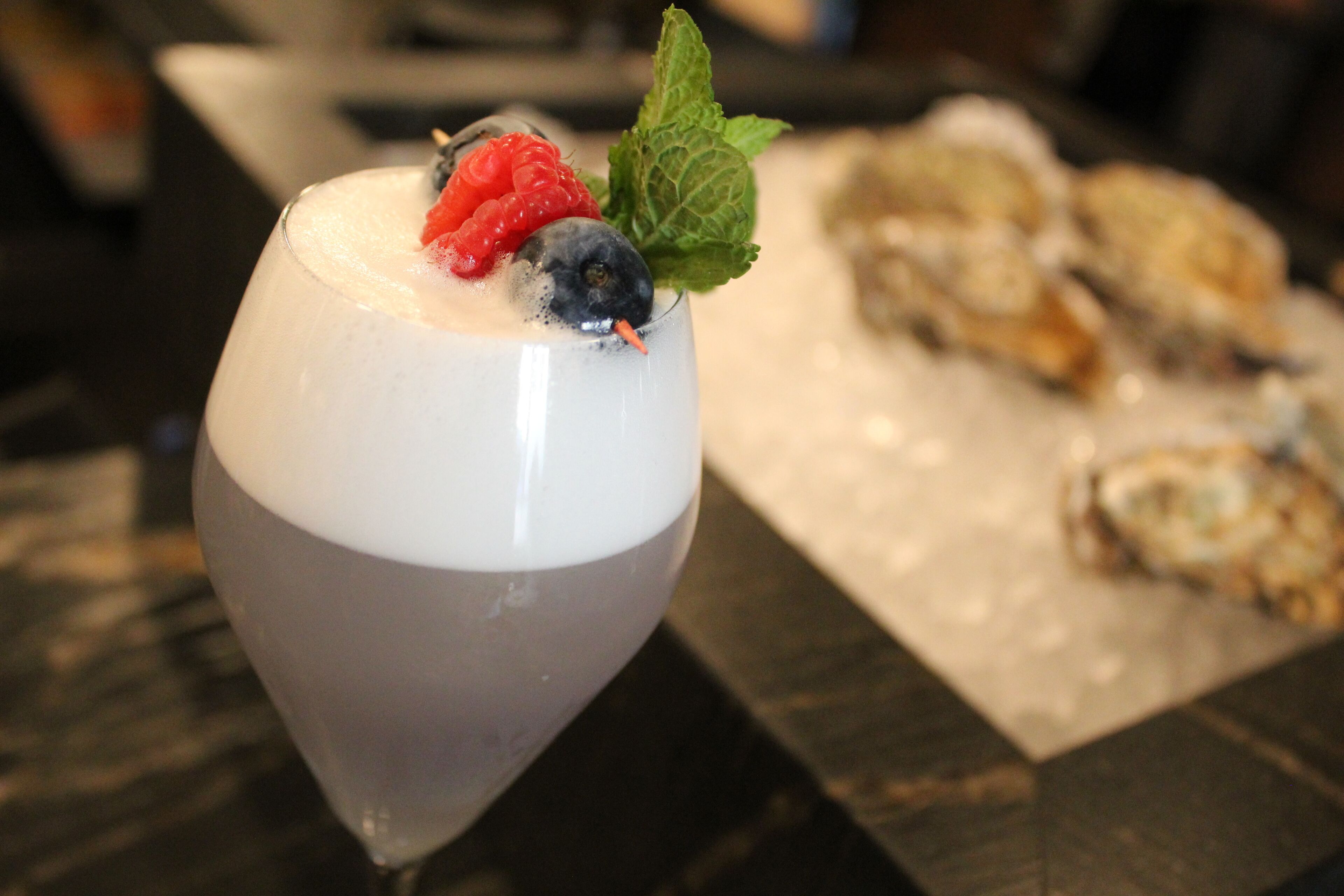 A dessert glass with a creamy dessert, garnished with a raspberry, blueberry, and mint leaf, on a table with a dessert tray in the background.