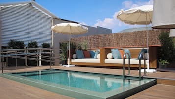 A swimming pool with a wooden deck, a white outdoor sofa with cushions, and two white umbrellas.