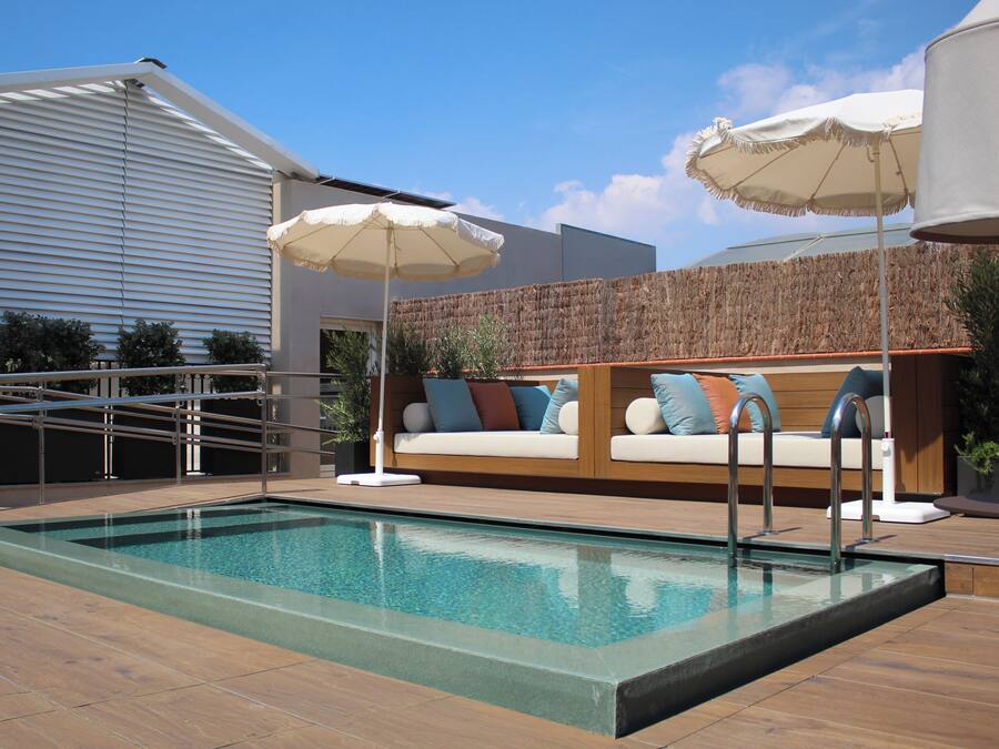 Una piscina con cubierta de madera, un sofá blanco para exteriores con cojines y dos sombrillas blancas.