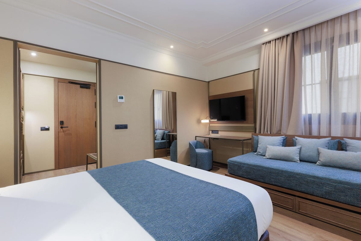 Quarto de hotel com uma cama grande, um sofá-cama, uma escrivaninha com cadeira, televisão e espelho.