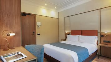 A hotel room with a large bed, a desk with a lamp, a chair, and a wooden wardrobe.