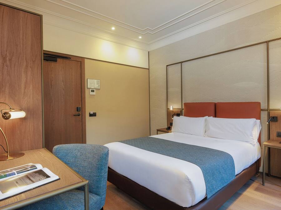 A hotel room with a large bed, a desk with a lamp, a chair, and a wooden wardrobe.