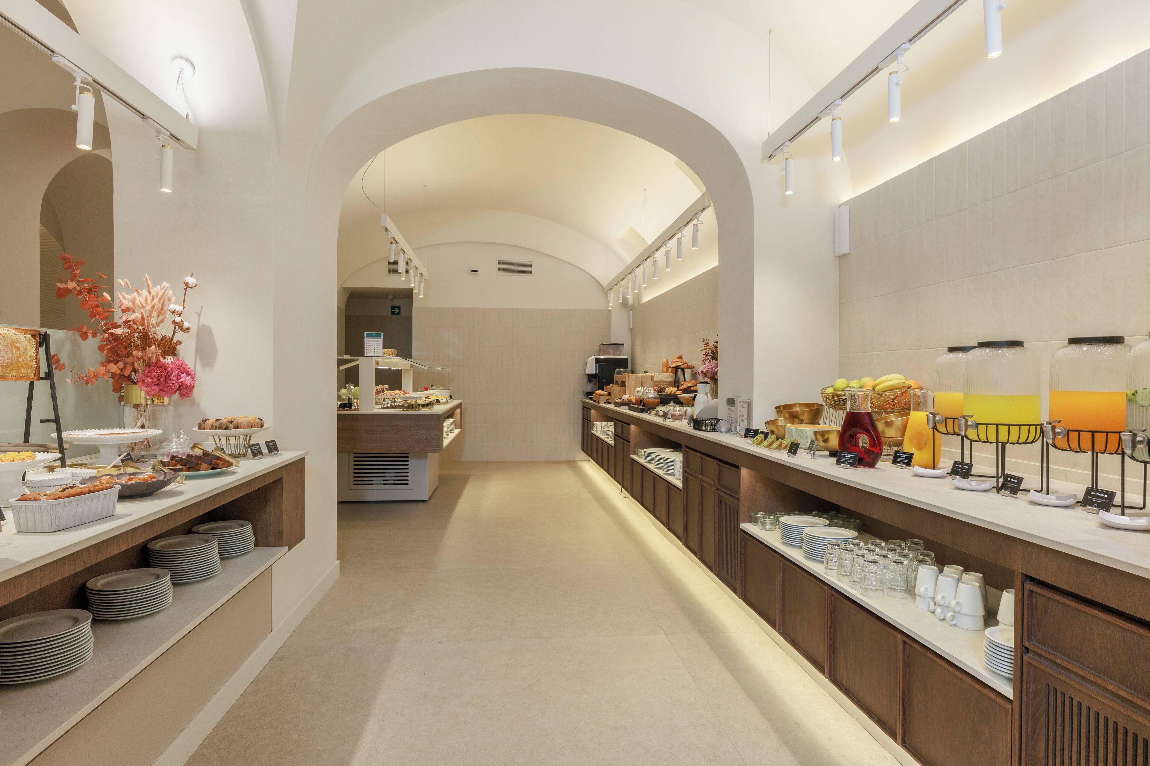 A spacious, well-lit hotel corridor with a modern design, featuring a long counter with various food and beverage options, and an arched ceiling.