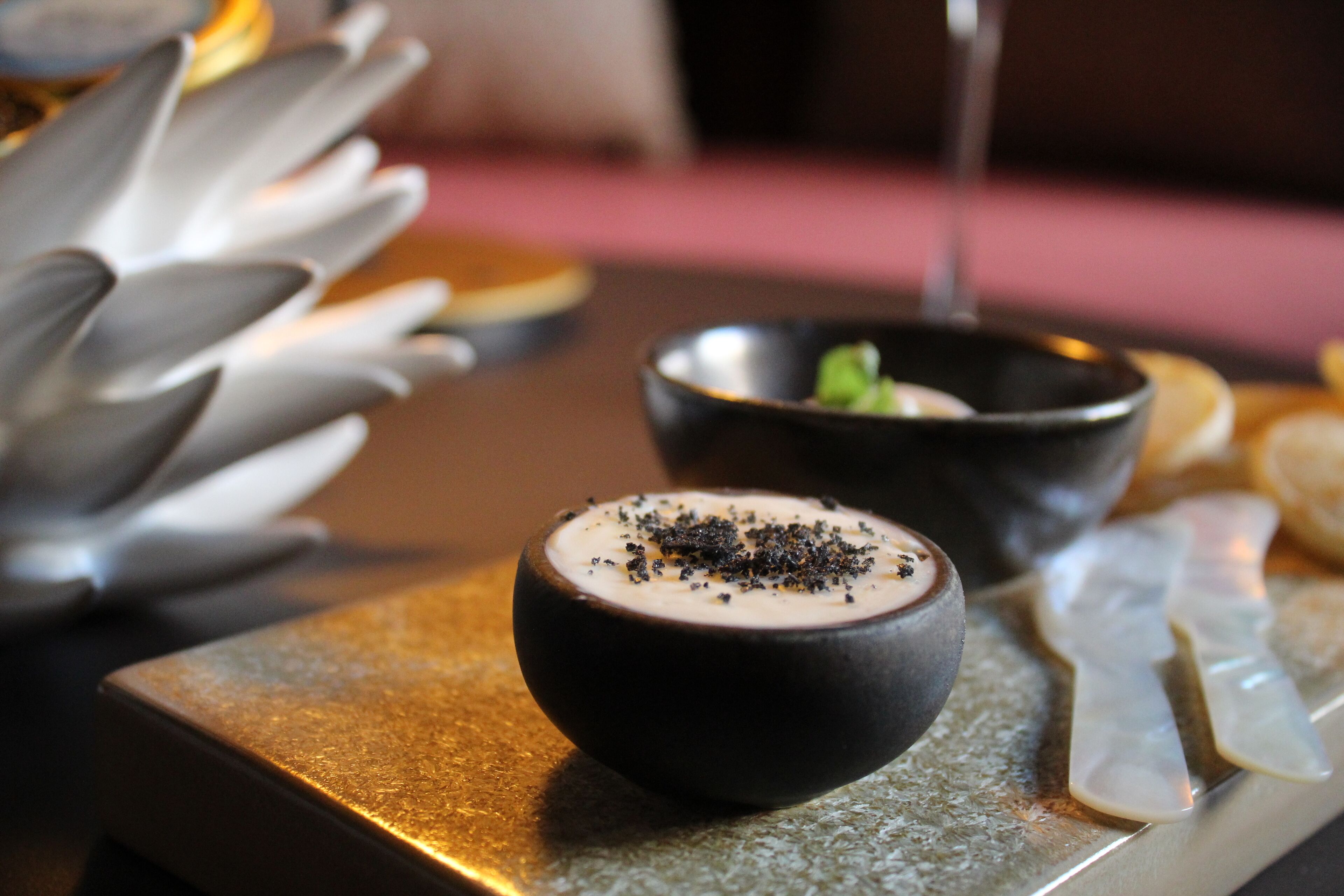 Presentación de postre con un mousse de chocolate en un bol negro, decorado con virutas de chocolate, sobre un plato de servicio texturizado.