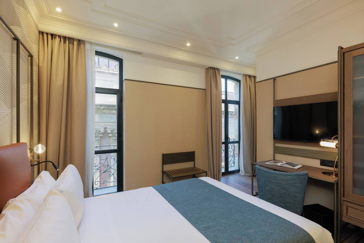 Quarto de hotel com uma cama grande, uma escrivaninha com cadeira, televisão e janela com cortinas.