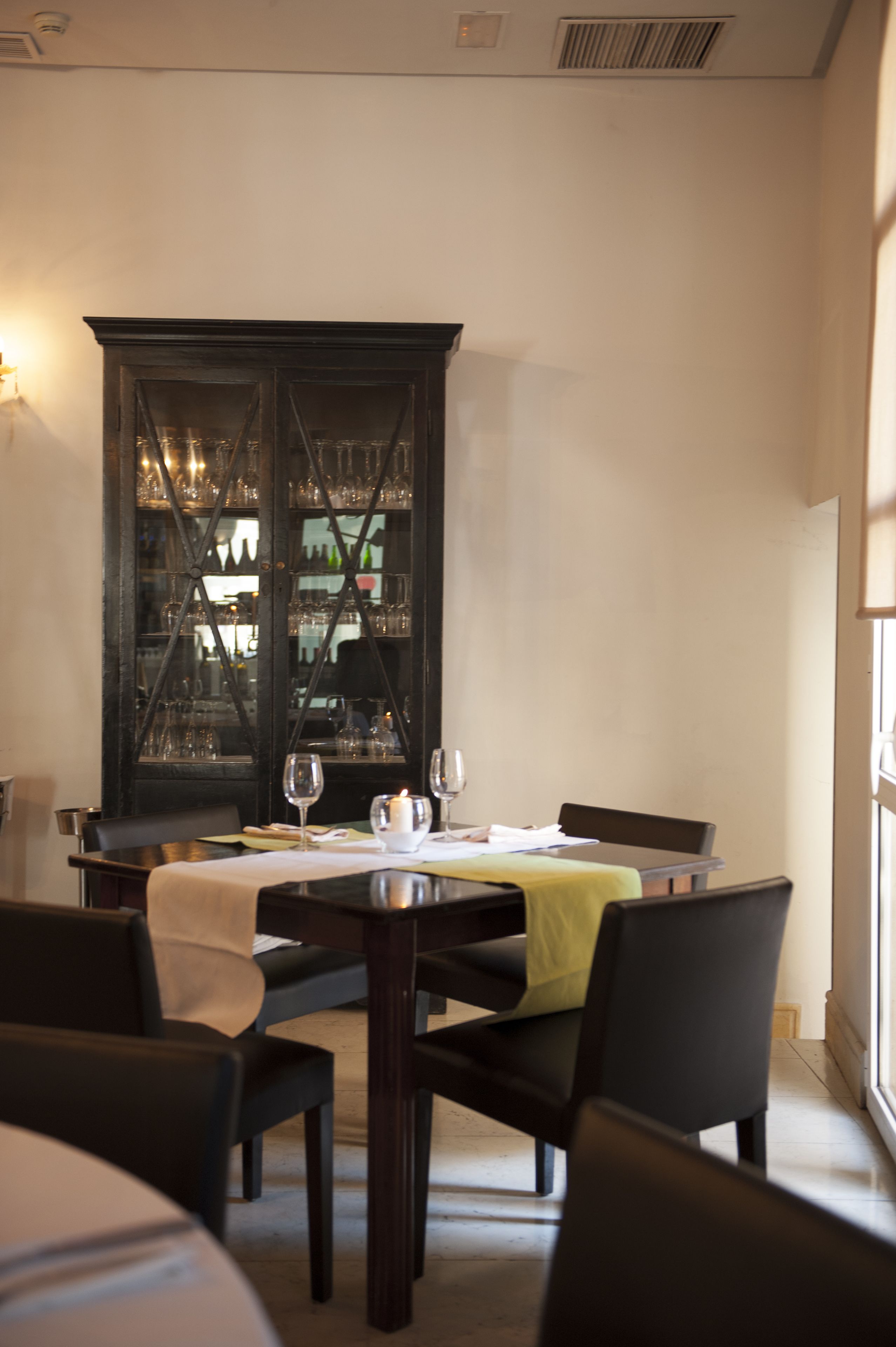 A dining area with a table set for two, a cabinet with glassware, and chairs.