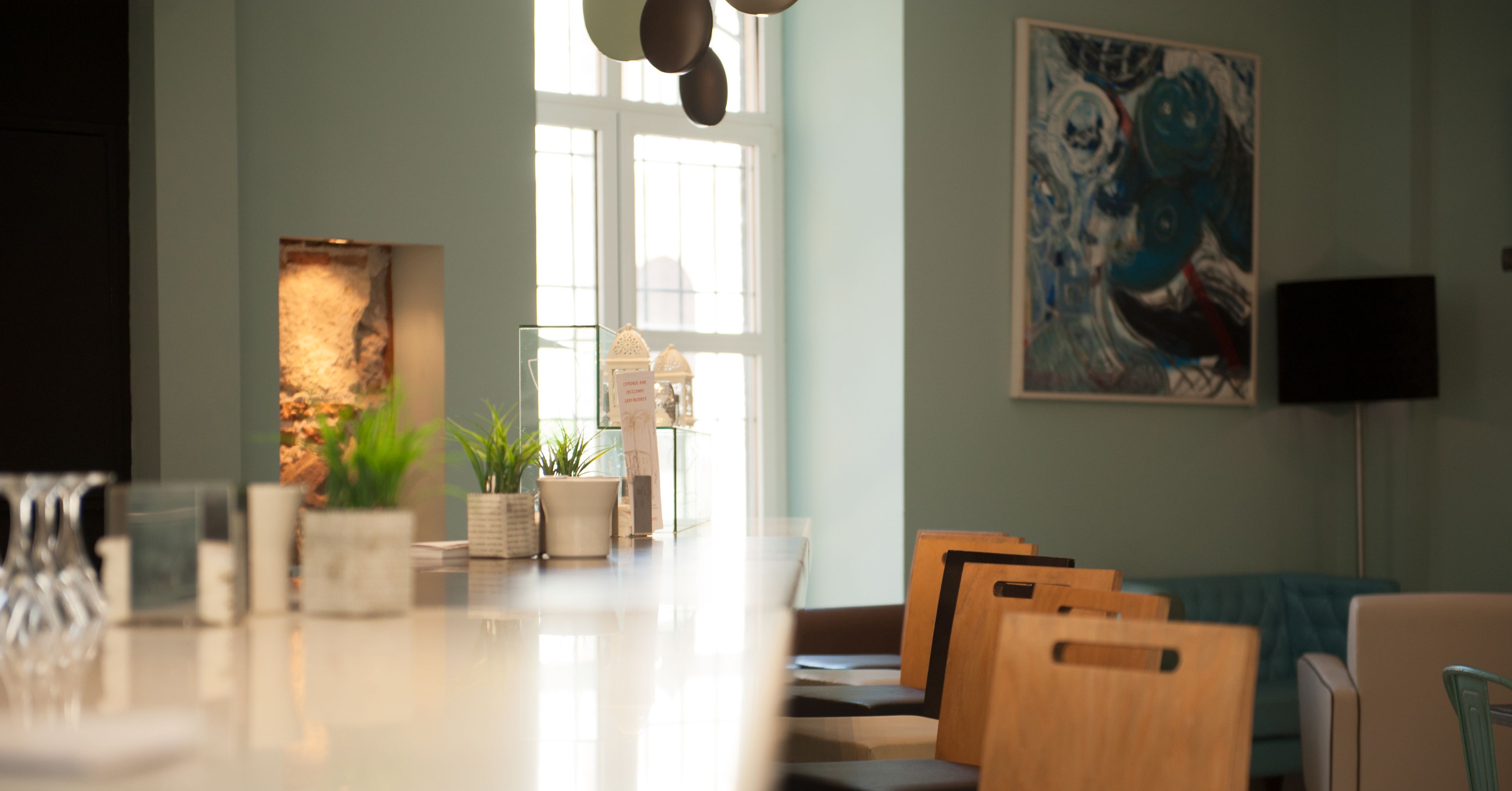 Un interior moderno con una larga mesa de comedor, sillas de madera, plantas en macetas, un cuadro abstracto grande y un televisor sobre un mueble bajo.