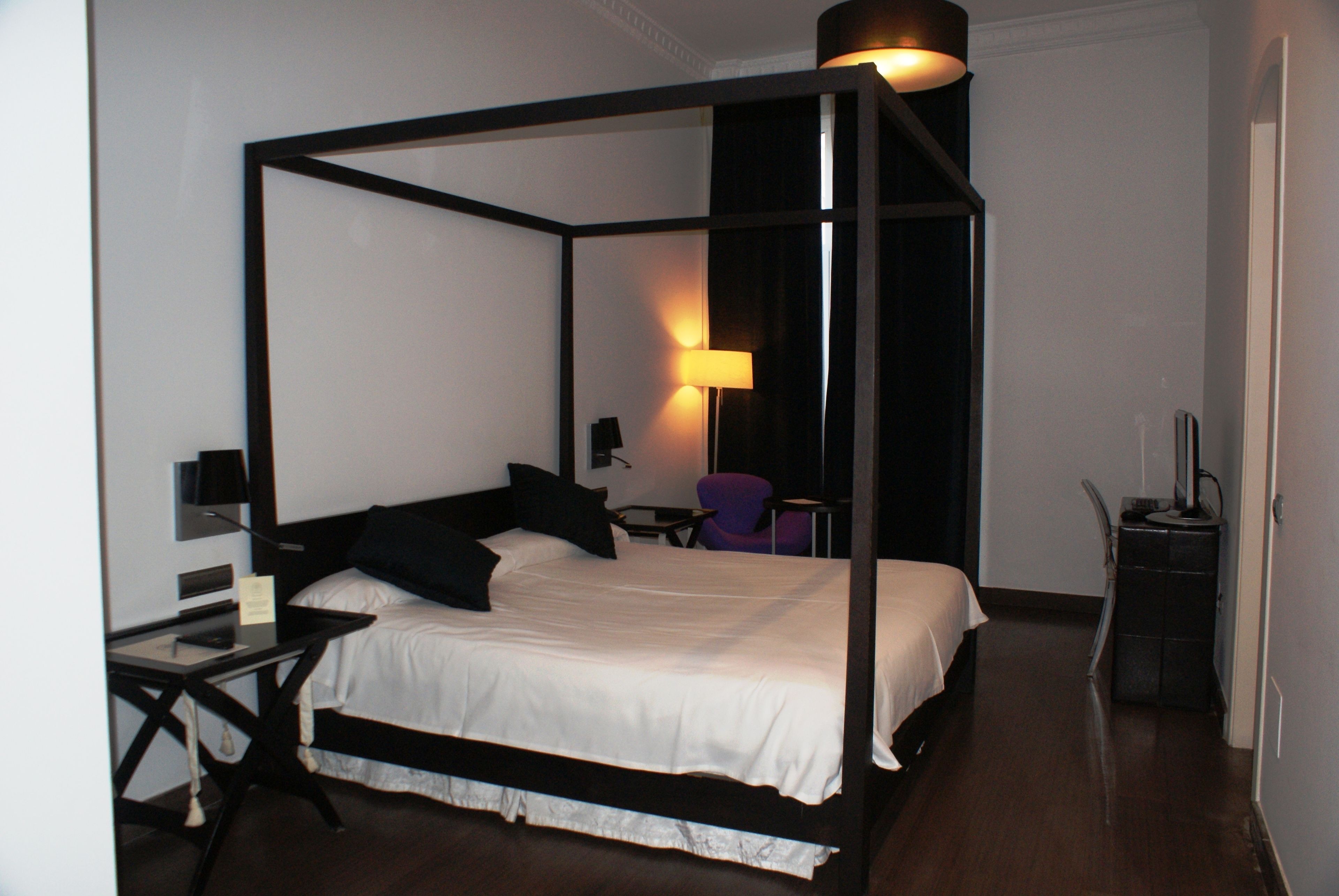 A four-poster bed with a black frame and white bedding, a desk with a chair, a lamp, and a small table.