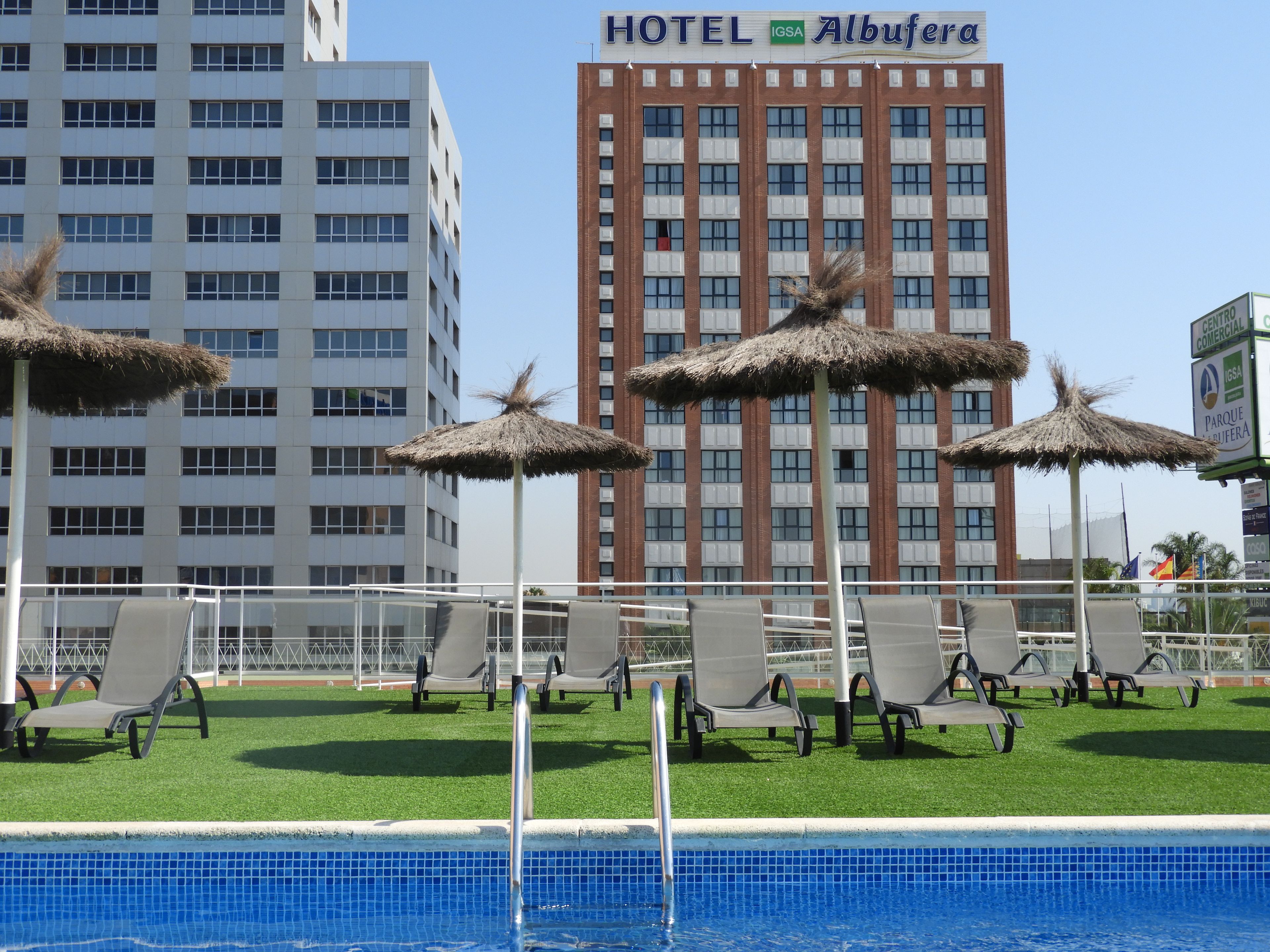 Une piscine d’hôtel entourée de chaises longues et de parasols en chaume, avec une eau d’un bleu limpide, et le bâtiment de l’hôtel en arrière-plan.
