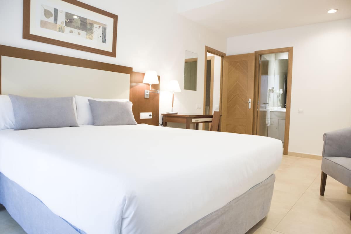 Habitación de hotel con una cama grande, un escritorio, una silla, una lámpara y un baño visible a través de una puerta abierta.