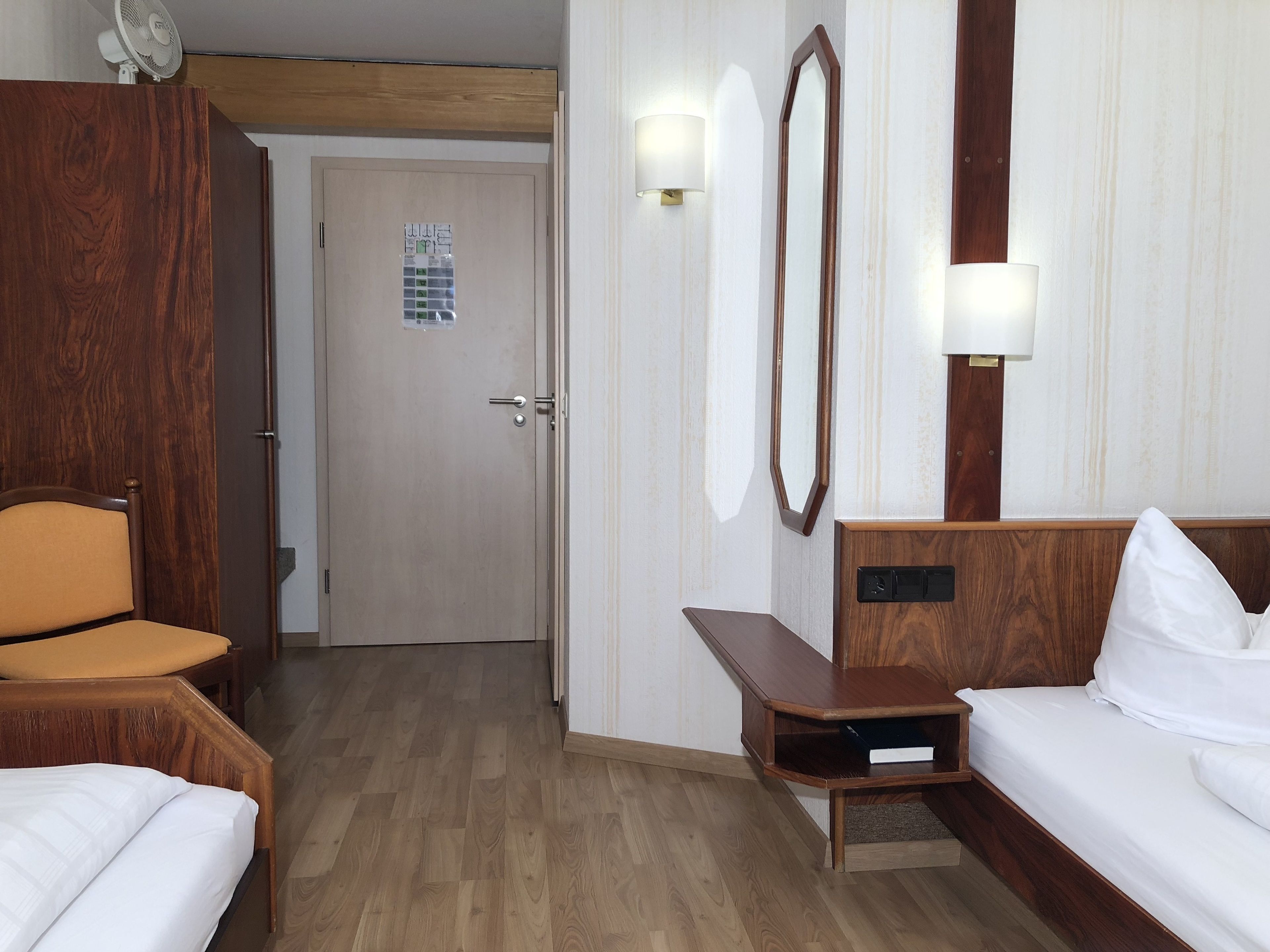 A hotel room with a wooden bed, a wooden nightstand, a wooden chair, a wooden desk, a mirror, and a door with a small window.
